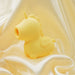 The Bean Blossom Unicorn Shaped Licking Tongue Vibrator in yellow, by Creative Conceptions, is a small, smooth figurine with closed eyes and a simple horn. It sits on a draped, glossy white fabric background, creating a soft and calming aesthetic. Crafted from premium silicone, its round shape and minimalist design give it a whimsical and friendly appearance.