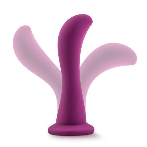 The Bellatrix 6.25 Inch G-Spot and Prostate Dildo in Plum Purple by Blush features an ergonomic silicone design with a smooth, curved shape and a flared base. Motion blurs in the image suggest flexibility and movement, making it perfect for g-spot or prostate stimulation. The white background ensures the product stands out clearly.