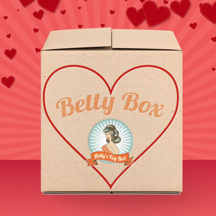 A cardboard pleasure box with a large red heart outline and "Betty's Penis Pleasures Mystery Box - Valentine's 2026 Edition" on the front. Set against a red background with hearts, it features a retro woman logo, hinting at surprises inside.