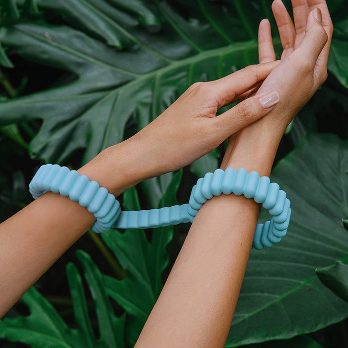A person's hands are gently connected by Biind Flexible Silicone Beginner Cuffs in blue, made of body-safe silicone, with large green leaves in the background.