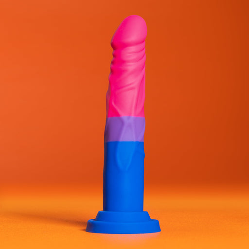The Bisexual Pride 7.5 Inch Silicone Suction Cup Dildo from Blush stands upright against an orange background. This multicolored, phallic-shaped object features a blue base, purple midsection, and pink tip, crafted from platinum cured silicone to proudly display the hues of the bisexual flag.