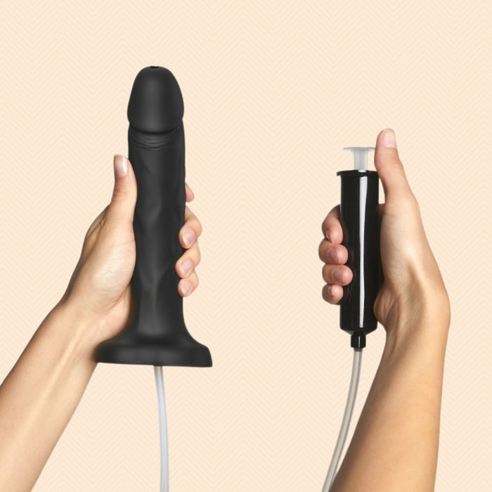A person holds the Strap-On-Me Realistic 7.5 Inch Silicone Squirting Dildo in black with its suction base, while grasping a bulb pump attached by hose, set against a light beige background.