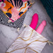 Two pink sex toys partially in a Blush Embrace White & Gold Toy Storage Bag rest on a marble surface. Above them are two other pouches with colorful abstract designs, all from Blush and featuring drawstring tie closures for secure sex toy storage. Made from natural reinforced cotton, these pouches ensure durability and style.