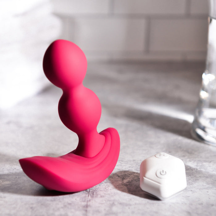 On a grey surface, the Evolved Novelties Bubble Butt Inflatable Vibrating Butt Plug with Remote—a pink, curved silicone adult toy featuring two bead-like segments and a wide base—rests next to its small white wireless remote control with buttons. The background showcases a bathroom setting with white tiles and a blurred towel.