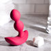 On a grey surface, the Evolved Novelties Bubble Butt Inflatable Vibrating Butt Plug with Remote—a pink, curved silicone adult toy featuring two bead-like segments and a wide base—rests next to its small white wireless remote control with buttons. The background showcases a bathroom setting with white tiles and a blurred towel.