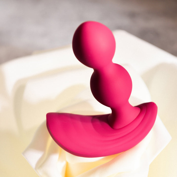 On a soft, light-colored fabric rests the Bubble Butt Inflatable Vibrating Butt Plug with Remote from Evolved Novelties. This red, dual-ball-shaped silicone object features a flared base for safety. The body-safe silicone butt plug has a smooth texture and is positioned upright, with the two spherical sections stacked vertically. The background is blurred and neutral in tone.