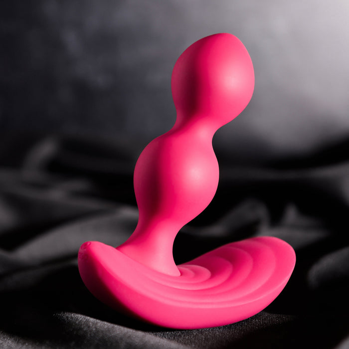 The Evolved Novelties Bubble Butt Inflatable Vibrating Butt Plug with Remote, a pink, body-safe silicone sex toy with a contoured, ergonomic design featuring two bulbous sections and a curved base, is placed against a dark, silky background.