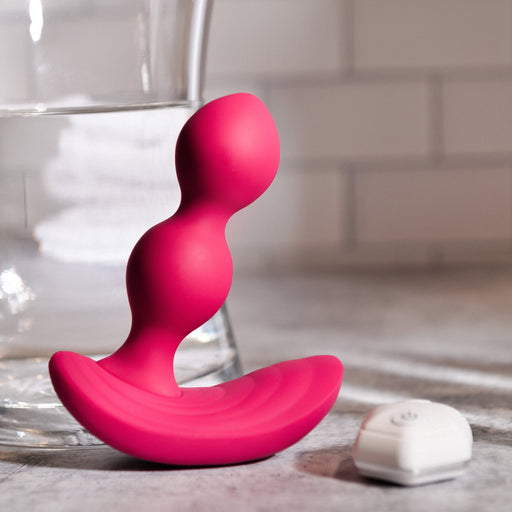 Close-up of a red, curved Evolved Novelties Bubble Butt Inflatable Vibrating Butt Plug with Remote featuring three bulbs, resting on a grey surface. A glass of water and a soft-focus tiled wall are in the background. Made from silicone, it has a sleek, modern design.