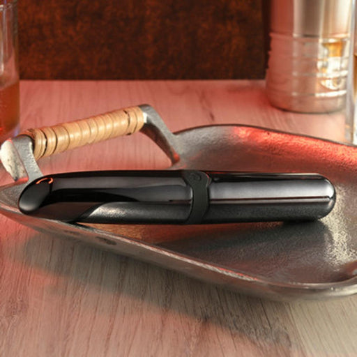 A sleek black Buzz Buddy Powerful Metal Vibrator with a curved design from Evolved Novelties sits on a textured metallic tray. A glass of amber liquid and a metallic container are partially visible in the background on a wooden surface. The scene is warmly lit, emphasizing the modern aesthetic of the vibrator.