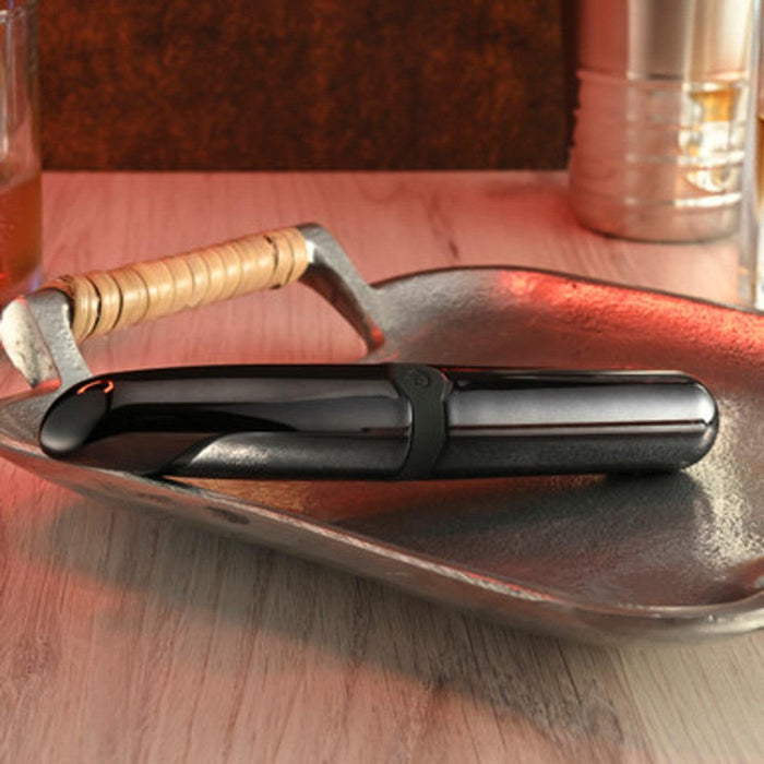 A sleek black Buzz Buddy Powerful Metal Vibrator with a curved design from Evolved Novelties sits on a textured metallic tray. A glass of amber liquid and a metallic container are partially visible in the background on a wooden surface. The scene is warmly lit, emphasizing the modern aesthetic of the vibrator.