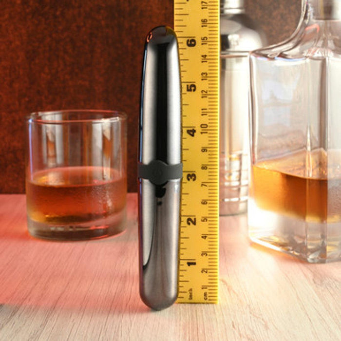 A sleek, metallic-colored Buzz Buddy Powerful Metal Vibrator from Evolved Novelties stands upright in front of a yellow measuring tape showing a height of about 5.5 inches. Behind the vibrator, there is a glass of amber liquid and a decanter filled with a similar liquid, placed on a wooden surface.