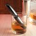 A glass of whiskey on a wooden table with a Buzz Buddy Powerful Metal Vibrator by Evolved Novelties inside. Nearby, a metal shaker and an open, partially filled whiskey bottle sit in the background, creating the perfect setup for an evening unwind.