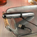 On a metal serving tray with a rope-wrapped handle, a sleek silver-colored electronic device from Evolved Novelties, the Buzz Buddy Powerful Metal Vibrator, sits connected to a USB charging cable. In the background, there’s a drink in a glass. This waterproof vibrator is perfect for any environment.