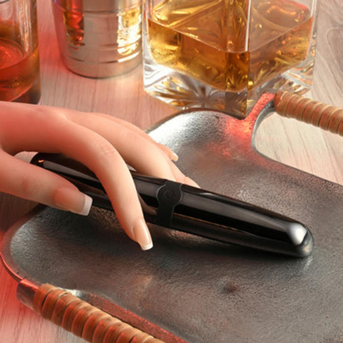 Close-up of a hand with long manicured nails holding a sleek and powerful Buzz Buddy Metal Vibrator from Evolved Novelties. The hand rests on a tray, exuding a beginner-friendly vibe, with a glass of amber liquid and another metallic container in the background on a wooden surface. The scene is warmly lit.