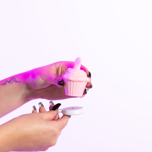 A person with long, painted nails holds the Cake Eater Clitoral Stimulator Tongue Vibrator in pink and its white round lid against a plain white background.