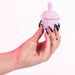 A hand with long black nails and rings holds the Cake Eater Clitoral Stimulator Tongue Vibrator in pink, a cupcake-shaped sex toy with a rounded tip for clitoral stimulation, against a light pink background.