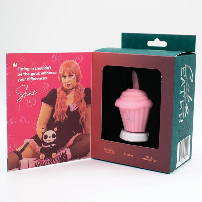 The Cake Eater Clitoral Stimulator Tongue Vibrator - Pink, shaped like a cupcake, is displayed in an open box featuring an image of a pink-haired person and plush toy. Inside, it says: "Fitting in shouldn't be the goal; embrace your differences.