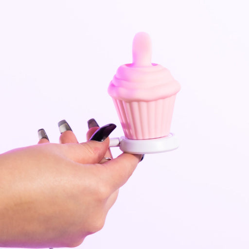 A hand with long, manicured nails holds the Cake Eater Clitoral Stimulator Tongue Vibrator in pink—shaped like a cupcake with a swirl top and white base—designed for clitoral pleasure, shown against a plain white background.