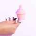 A hand with long, manicured nails holds the Cake Eater Clitoral Stimulator Tongue Vibrator in pink—shaped like a cupcake with a swirl top and white base—designed for clitoral pleasure, shown against a plain white background.