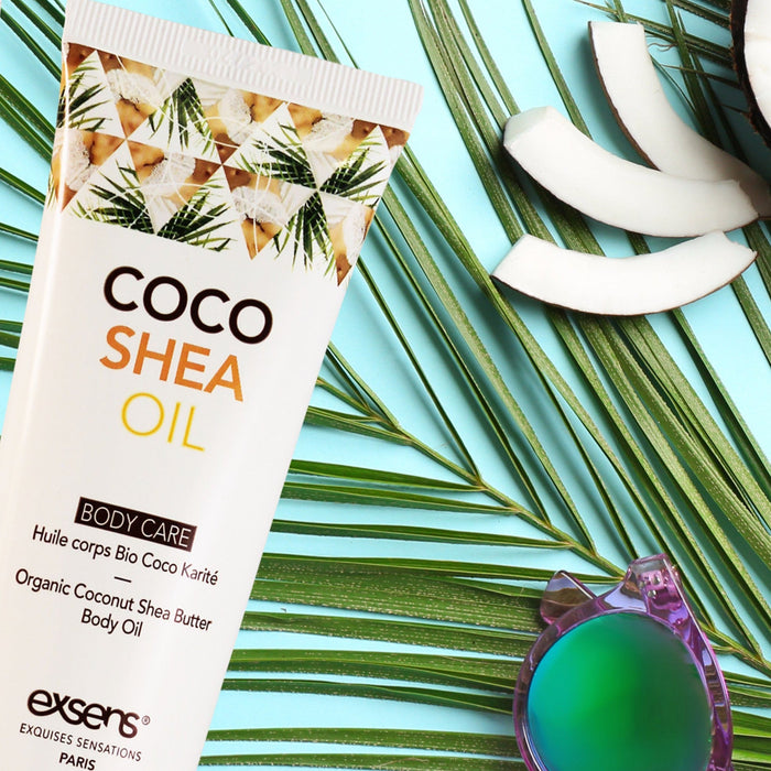 A tube of Exsens Coco Shea Organic Body Oil & Intimate Moisturizer, perfect as a massage oil and dry skin solution, rests on palm leaves against a light blue background. The tube displays tropical coconut graphics and the text "Organic Coconut Shea Butter Body Oil" in multiple languages. Nearby, pieces of coconut and a green round glass object complete the scene.