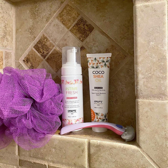 A shower niche featuring a purple bath pouf, a bottle of Intime Fresh body wash, a tube of Exsens Coco Shea Organic Body Oil & Intimate Moisturizer for dry skin solutions, and a pink-handled razor. The background consists of beige and brown textured tiles.