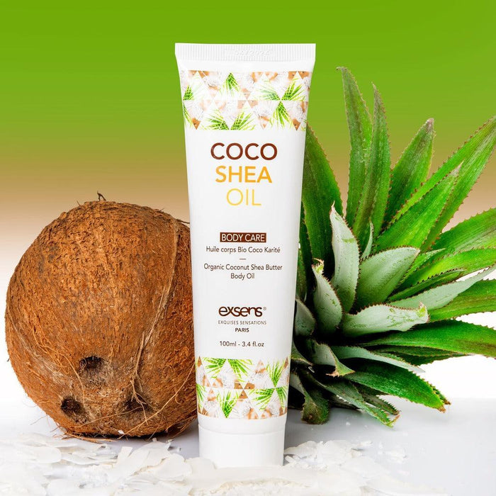 A bottle of Exsens Coco Shea Organic Body Oil & Intimate Moisturizer stands upright on a white surface scattered with coconut flakes. The background features a whole coconut and an aloe vera plant against a green gradient backdrop, highlighting its effectiveness as an intimate moisturizer.