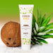 A bottle of Exsens Coco Shea Organic Body Oil & Intimate Moisturizer stands upright on a white surface scattered with coconut flakes. The background features a whole coconut and an aloe vera plant against a green gradient backdrop, highlighting its effectiveness as an intimate moisturizer.