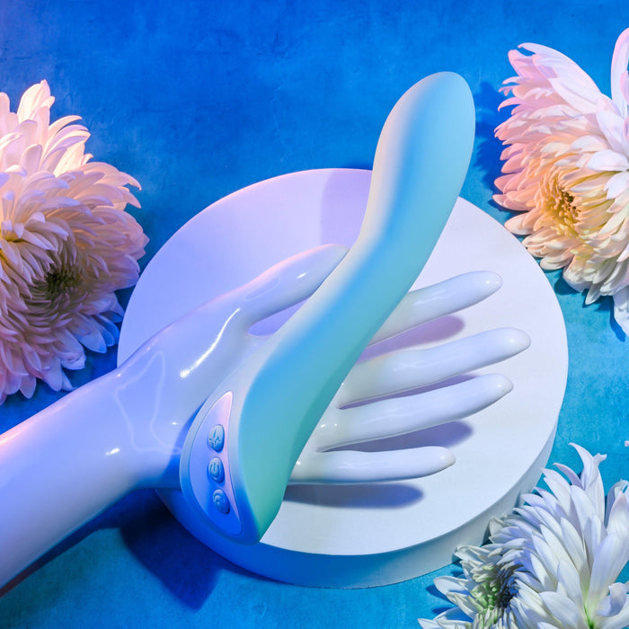 A teal "Come with Me G-Spot Vibrator with Come Hither Motion" from Evolved Novelties, featuring control buttons on its side, rests on a white mannequin hand perched on a white round pedestal. The setup is surrounded by white flowers, all set against a vibrant blue background.