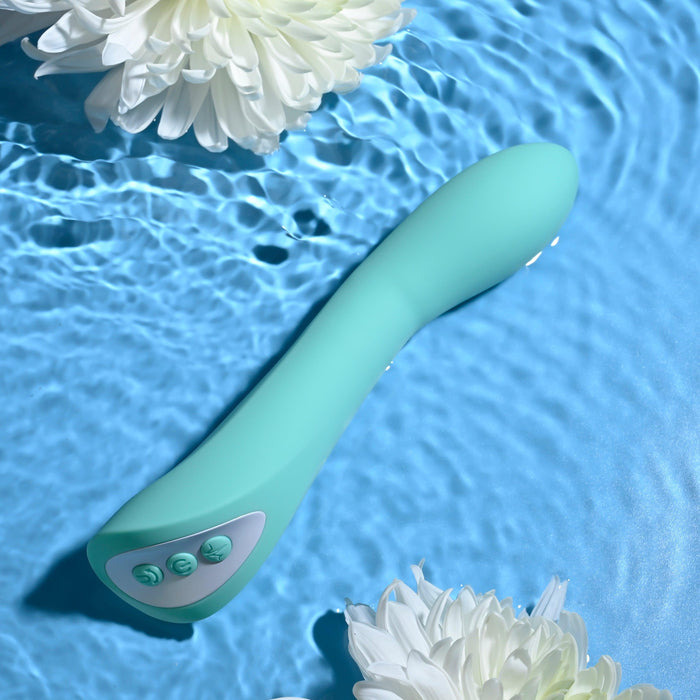 The Evolved Novelties Come with Me G-Spot Vibrator with Come Hither Motion in teal, featuring three buttons, is submerged in clear blue water. Two white flowers float near the water submersible device, creating a serene and refreshing ambiance.