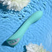 The Evolved Novelties Come with Me G-Spot Vibrator with Come Hither Motion in teal, featuring three buttons, is submerged in clear blue water. Two white flowers float near the water submersible device, creating a serene and refreshing ambiance.