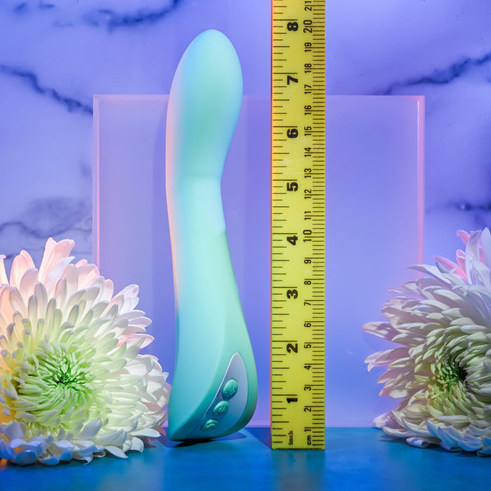 A teal-colored Evolved Novelties Come with Me G-Spot Vibrator with Come Hither Motion stands upright next to a yellow measuring tape, showcasing its length as approximately 7.5 inches. Featuring a purple gradient and marble pattern in the background, accompanied by white flowers on either side, this sleek g-spot vibrator is also water submersible for versatile pleasure.
