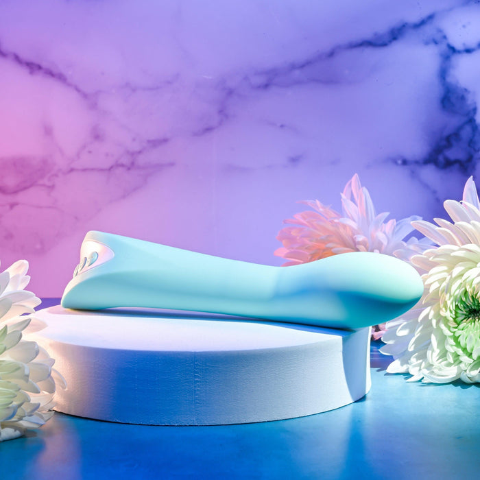 The *Come with Me G-Spot Vibrator with Come Hither Motion* by *Evolved Novelties* in teal rests on a white pedestal, set against a vibrant backdrop blending purple and blue tones with a marble-like texture. The scene is adorned with white flowers, creating a soft and elegant ambiance.