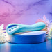 The Come with Me G-Spot Vibrator with Come Hither Motion in teal from Evolved Novelties, featuring a sleek curved design and three buttons on its side, is displayed on a round, white platform. The background showcases a pastel gradient of purple and blue adorned with white flowers on either side, creating a soft, soothing ambiance.
