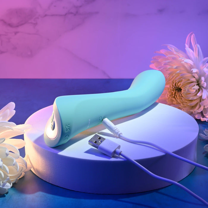 The Come with Me G-Spot Vibrator with Come Hither Motion in teal, from Evolved Novelties, rests on a circular white platform. A USB charging cable is placed beside it. The background features a soft purple and blue gradient with white flowers on either side of the g-spot massager.