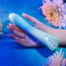 A hand holds the "Come with Me G-Spot Vibrator with Come Hither Motion - Teal" by Evolved Novelties against a blue surface. Soft pink flowers are artfully arranged around the scene, enhancing the delicate and serene atmosphere ideal for g-spot stimulation.