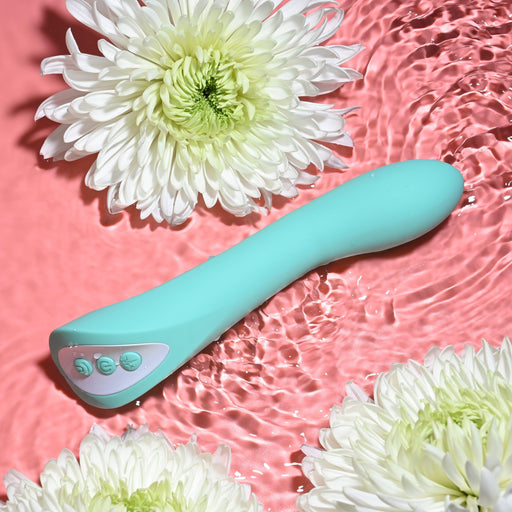 The "Come with Me G-Spot Vibrator with Come Hither Motion" by Evolved Novelties, in a calming teal color and equipped with three buttons, is partially submerged in water. Large white chrysanthemums surround it, contributing to its soft and soothing aesthetic. The pink background adds a striking contrast, while the ripples in the water enhance the allure of this g-spot pleasure device.