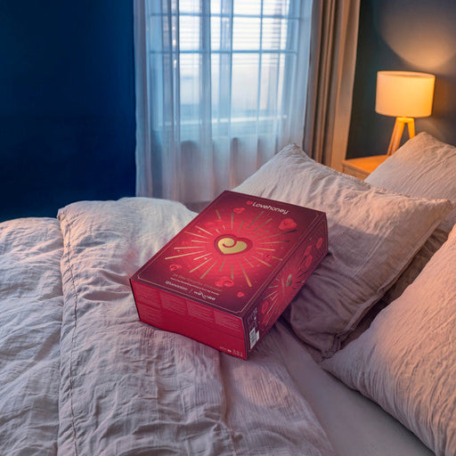 A red rectangular product box with a gold heart design sits on a bed with white and beige pillows in a bedroom with blue walls, a window with sheer curtains, and a lit bedside lamp.