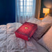 A red rectangular product box with a gold heart design sits on a bed with white and beige pillows in a bedroom with blue walls, a window with sheer curtains, and a lit bedside lamp.