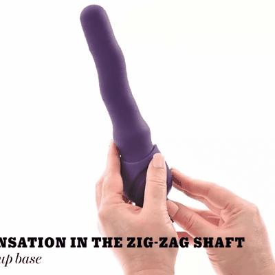 A person holds the Wiggle It Silicone Wiggling Vibrator with Suction Cup Base, which is purple and features a zig-zag shaped shaft. The bottom text partially reads, “SENSATION IN THE ZIG-ZAG SHAFT... up base.”.