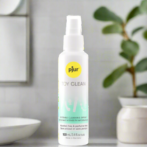A bottle of Pjur Toy Foam Gentle Cleaner for Sex Toys, labeled alcohol-free and perfume-free, is on a surface with a blurred background showing a plant and decor. Safe for all sex toys, it's an effective hygiene cleaning foam spray.