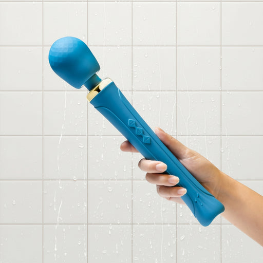 A hand holds the Dianna Rechargeable Rumbly Waterproof Wand Vibrator by Blush, showcasing its teal-colored, luxurious design with a textured ball-shaped head. The massager features a flexible neck, three buttons on the handle, and elegant gold accents near the head. The background is white.