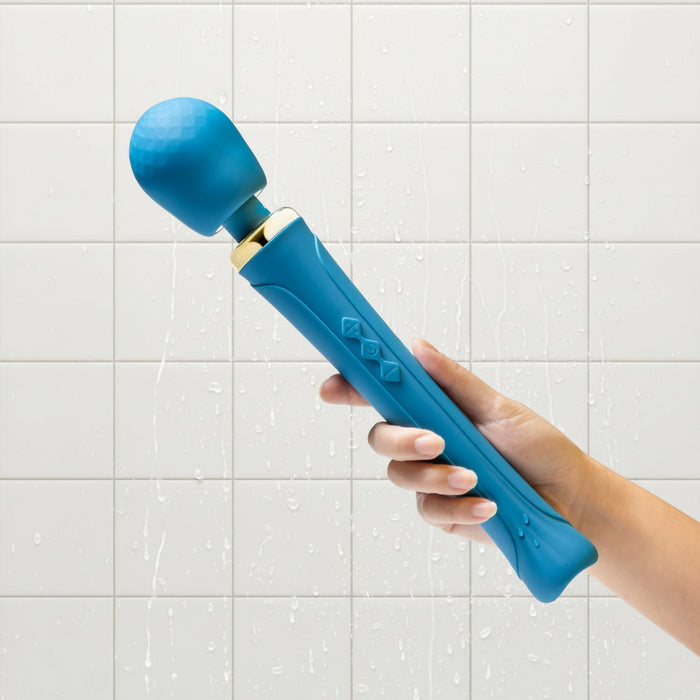 A hand holds the Dianna Rechargeable Rumbly Waterproof Wand Vibrator by Blush, showcasing its teal-colored, luxurious design with a textured ball-shaped head. The massager features a flexible neck, three buttons on the handle, and elegant gold accents near the head. The background is white.