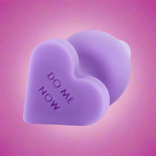 The Do Me Now Naughty Candy Heart Purple Butt Plug by Blush is heart-shaped with a slightly bulbous, rounded end and the words "DO ME NOW" etched onto its silicone surface. Positioned on a white background and slightly tilted to the side, it is ideal for anal beginners.