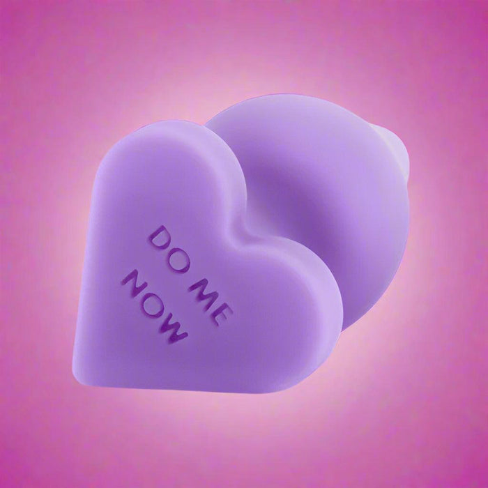 The Do Me Now Naughty Candy Heart Purple Butt Plug by Blush is heart-shaped with a slightly bulbous, rounded end and the words "DO ME NOW" etched onto its silicone surface. Positioned on a white background and slightly tilted to the side, it is ideal for anal beginners.