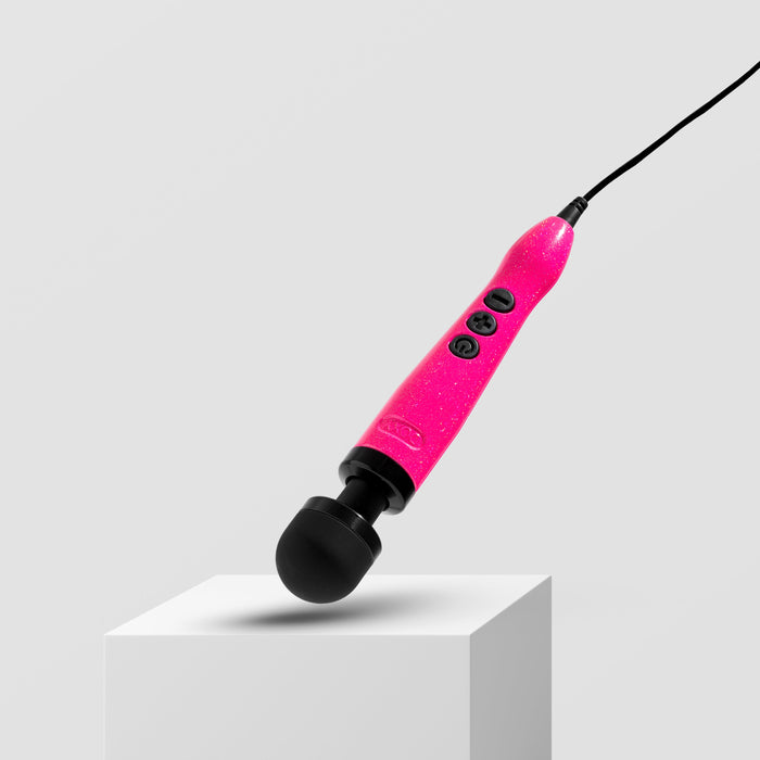 A bright pink handheld massage wand, named the Doxy Die Cast 3 Compact Wand Vibrator Hot Pink by Doxy, featuring black controls and a black rounded head, is positioned diagonally on a white box against a plain light gray background. This compact powerhouse is plugged in, with a cable extending from the end.