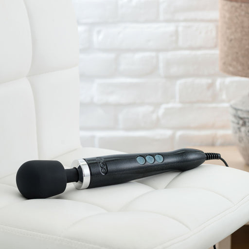 A sleek black Doxy Die Cast Extra Powerful Wand Vibrator with three buttons on the handle is placed on a white cushioned chair. The background features a white brick wall and part of a wooden table with a metallic lamp base, perfectly setting the scene for deep rumbly vibrations from your Doxy.