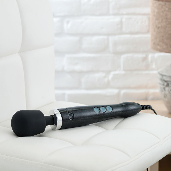 A sleek black Doxy Die Cast Extra Powerful Wand Vibrator with three buttons on the handle is placed on a white cushioned chair. The background features a white brick wall and part of a wooden table with a metallic lamp base, perfectly setting the scene for deep rumbly vibrations from your Doxy.