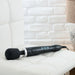 A sleek black Doxy Die Cast Extra Powerful Wand Vibrator with three buttons on the handle is placed on a white cushioned chair. The background features a white brick wall and part of a wooden table with a metallic lamp base, perfectly setting the scene for deep rumbly vibrations from your Doxy.