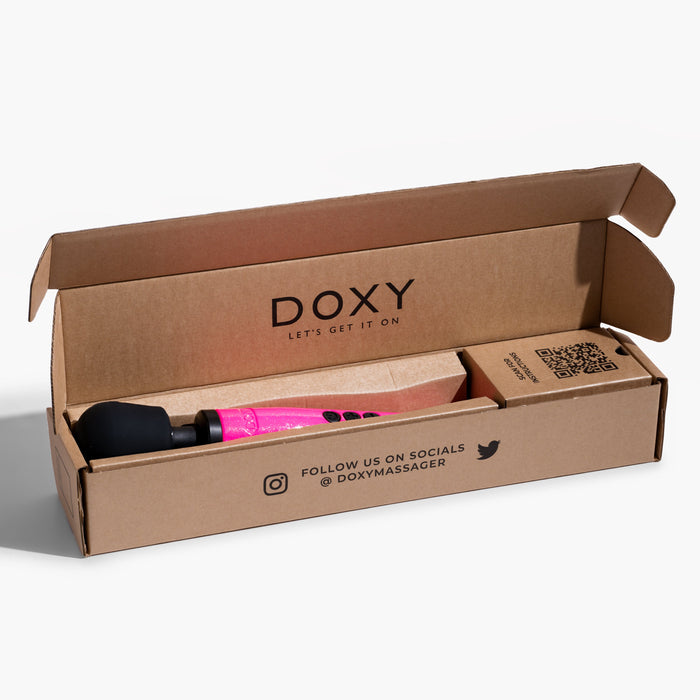 A cardboard box branded with "DOXY" and the tagline "LET'S GET IT ON" contains a Doxy Die Cast Extra Powerful Wand Vibrator in hot pink, featuring a black head that delivers deep vibrations. The box also includes printed social media handles and a QR code for more information.