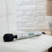 The Doxy Die Cast Extra Powerful Wand Vibrator – Silver, featuring a silver handle, black head, and three green control buttons, rests elegantly on a white cushioned chair. The pristine white brick wall in the background accentuates the scene.
